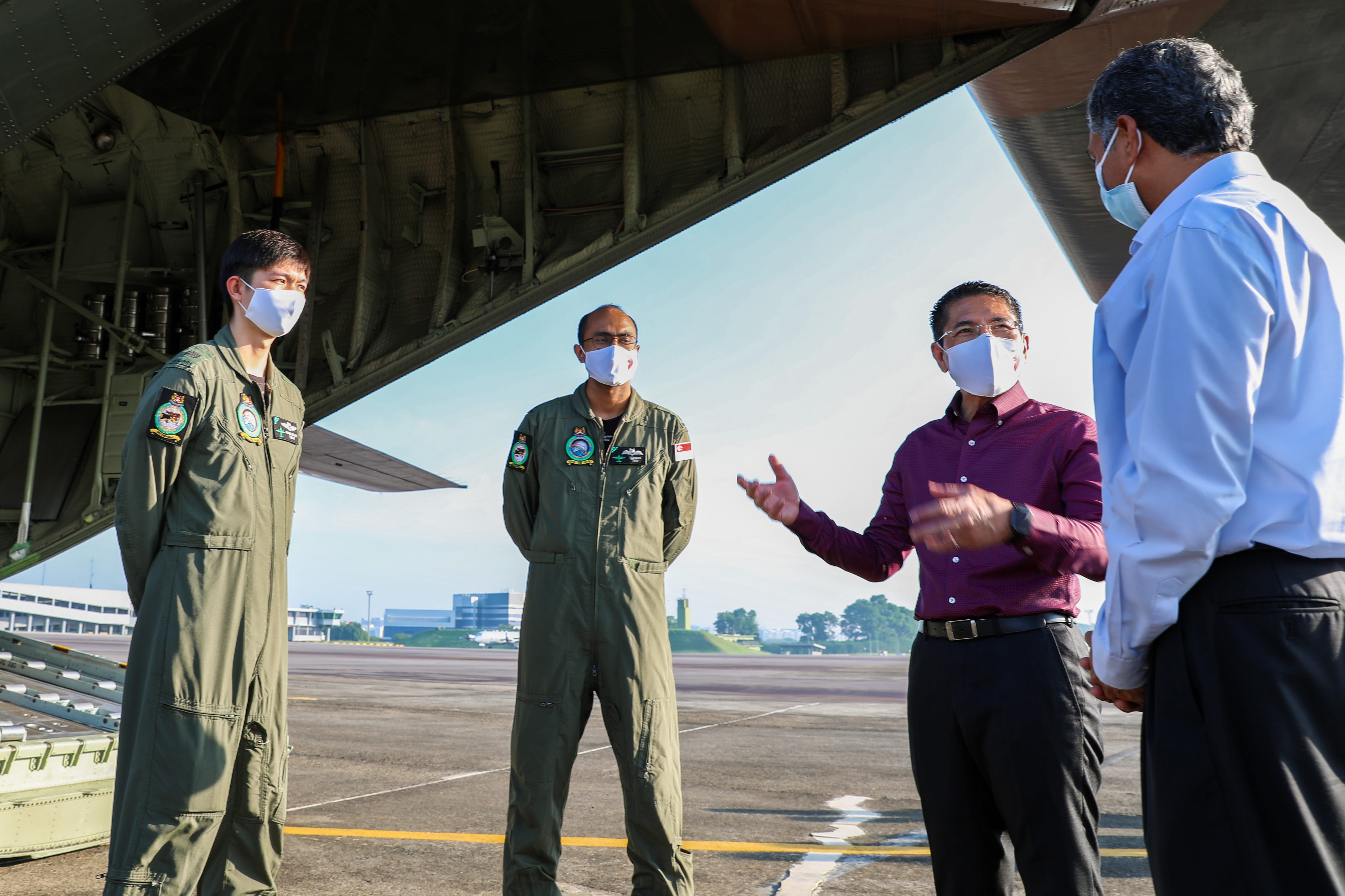 Four people wearing masks stand under an aircraft wing. Two are in military flight suits.